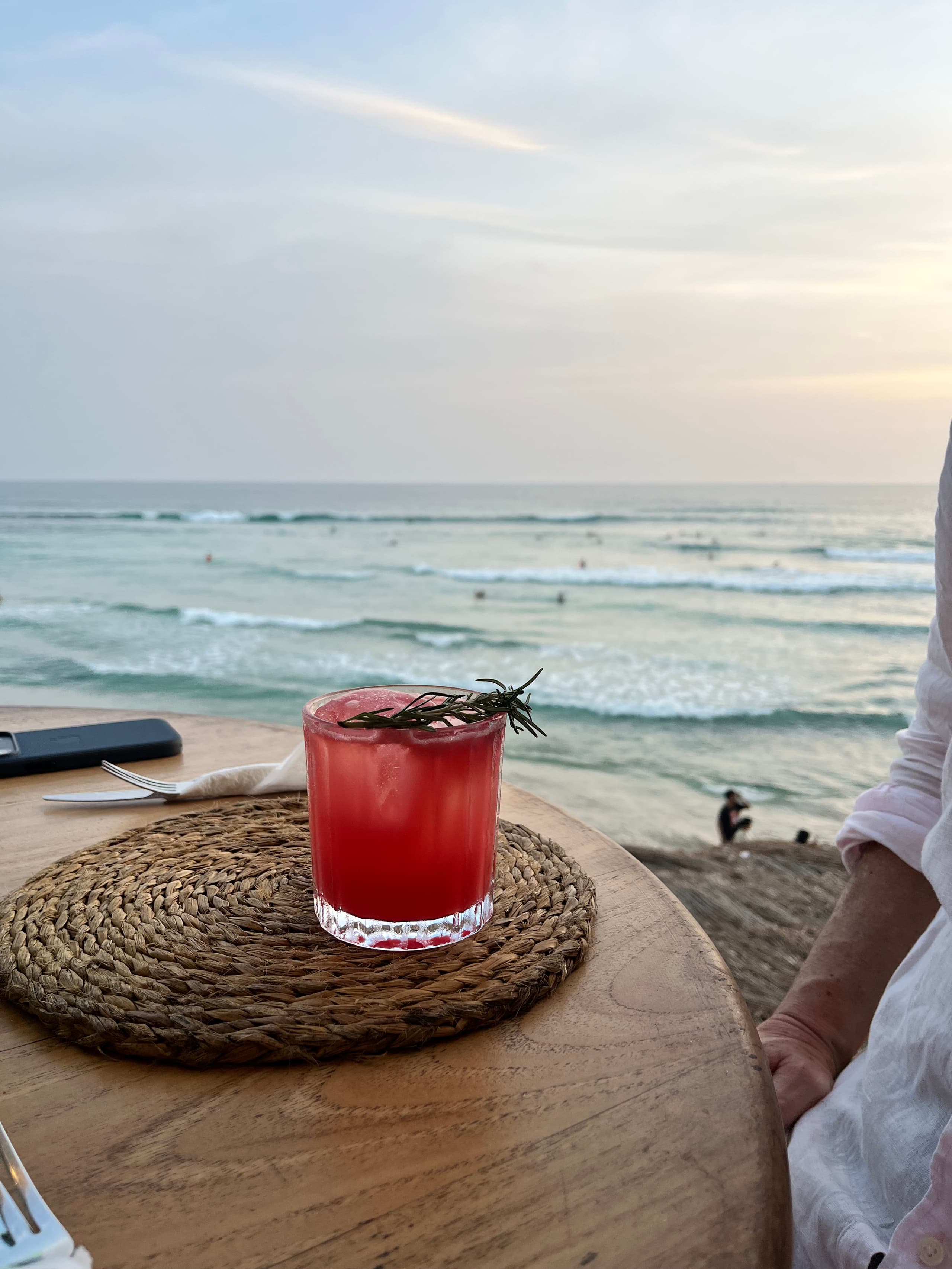 Red drink with rosemary on a table above the surf at sunset